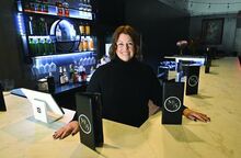 A woman in black stands at a bar surrounded by wine bottles, wine glasses and menus