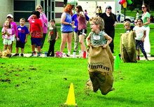 A small child with glasses and a big smile is airborne in a burlap sack on a very green grass field.