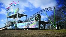 A green rollercoaster track with upholstered cars in the loading area and purple, white and blue flags flying, sits in a field.