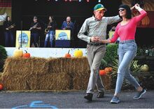 A beaming woman dances with a man in a baseball cap as a band plays in the background.