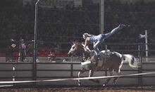 A woman balances on one leg on the back of a horse as it gallops around an arena.